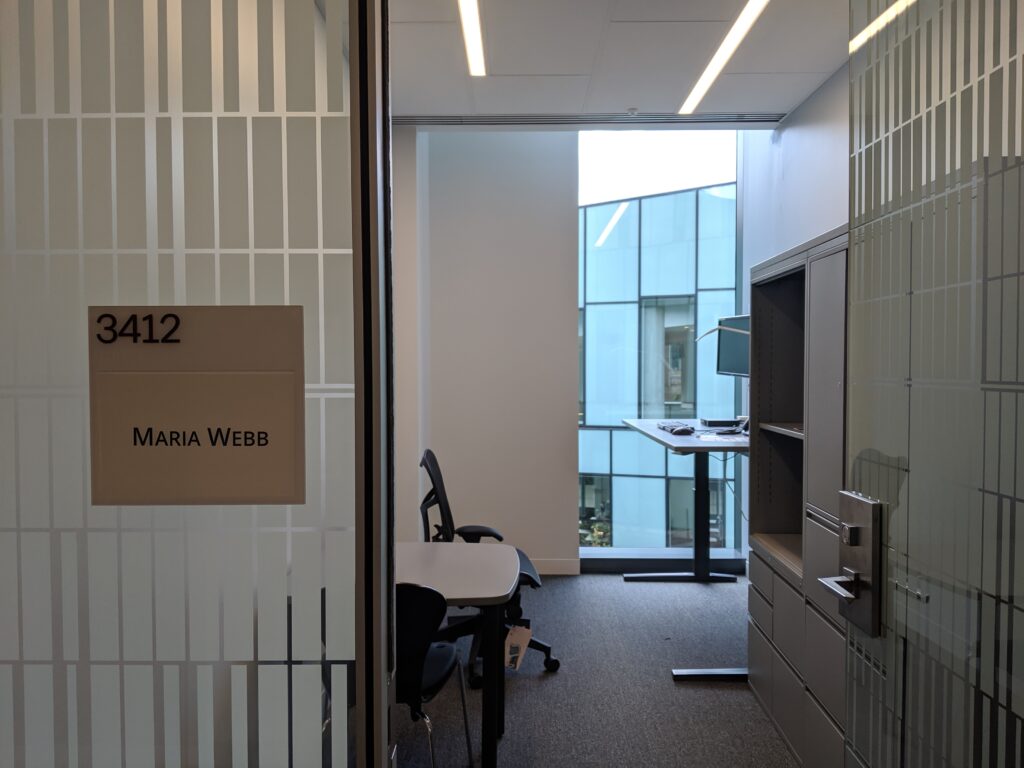 Picture of an empty college professor's office; you can see a small table, office chair, and standing desk with monitor in the background, and filing cabinet near the door. There is a big window in the back of the room.